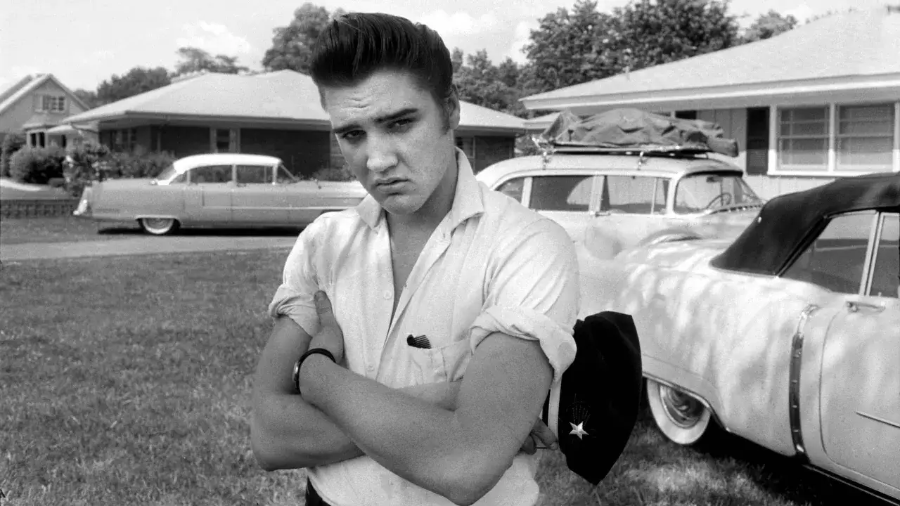 Elvis Presley, mit verschr&auml;nkten Armen und einem ernsten Blick, steht vor seinen Autos. Sein Verm&ouml;gen, der Elvis Presley net worth, war schon damals beeindruckend.