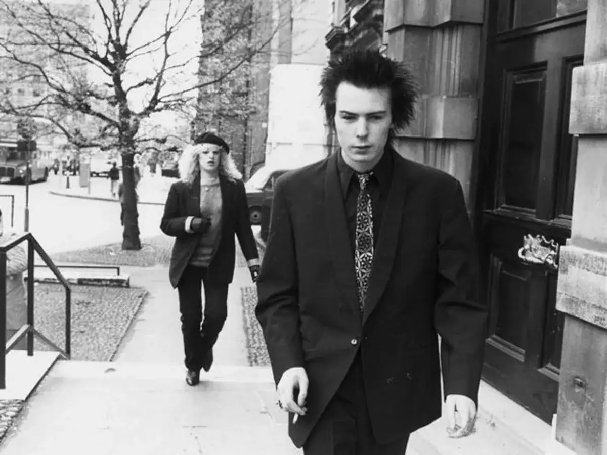 Sid Vicious und Nancy Spungen gehen eine Treppe hinunter. Sid, mit zerzaustem Haar und Anzug, scheint in Gedanken versunken, vielleicht erfüllt er seine sid wishes.