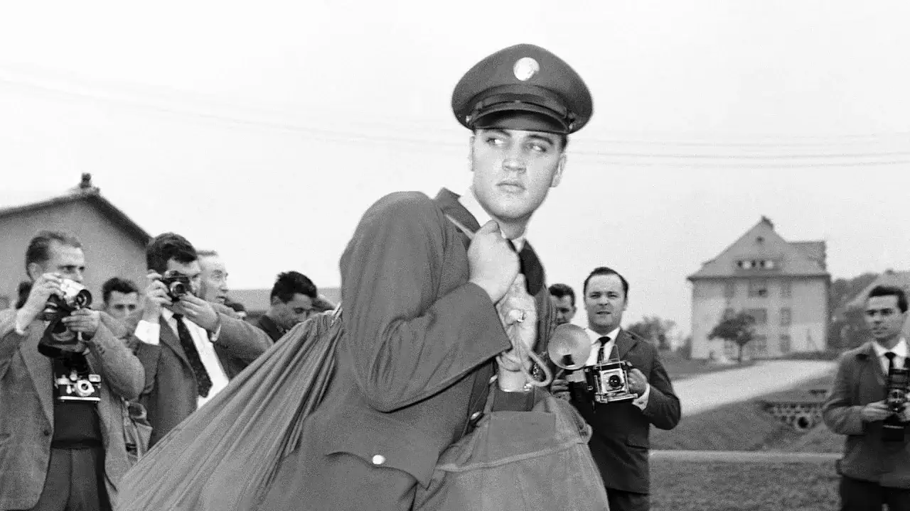 Elvis Presley im Armeedienst, umgeben von Paparazzi. Er blickt nachdenklich, während Fotografen mit Kameras auf ihn richten.