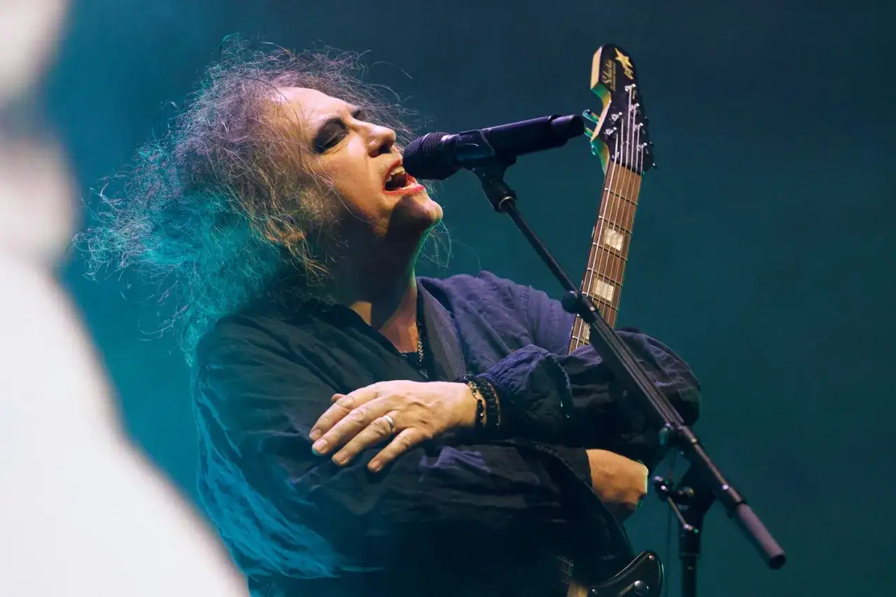 Robert Smith von The Cure singt mit geschlossenen Augen und zerzaustem Haar ins Mikrofon, während er seine Gitarre hält.