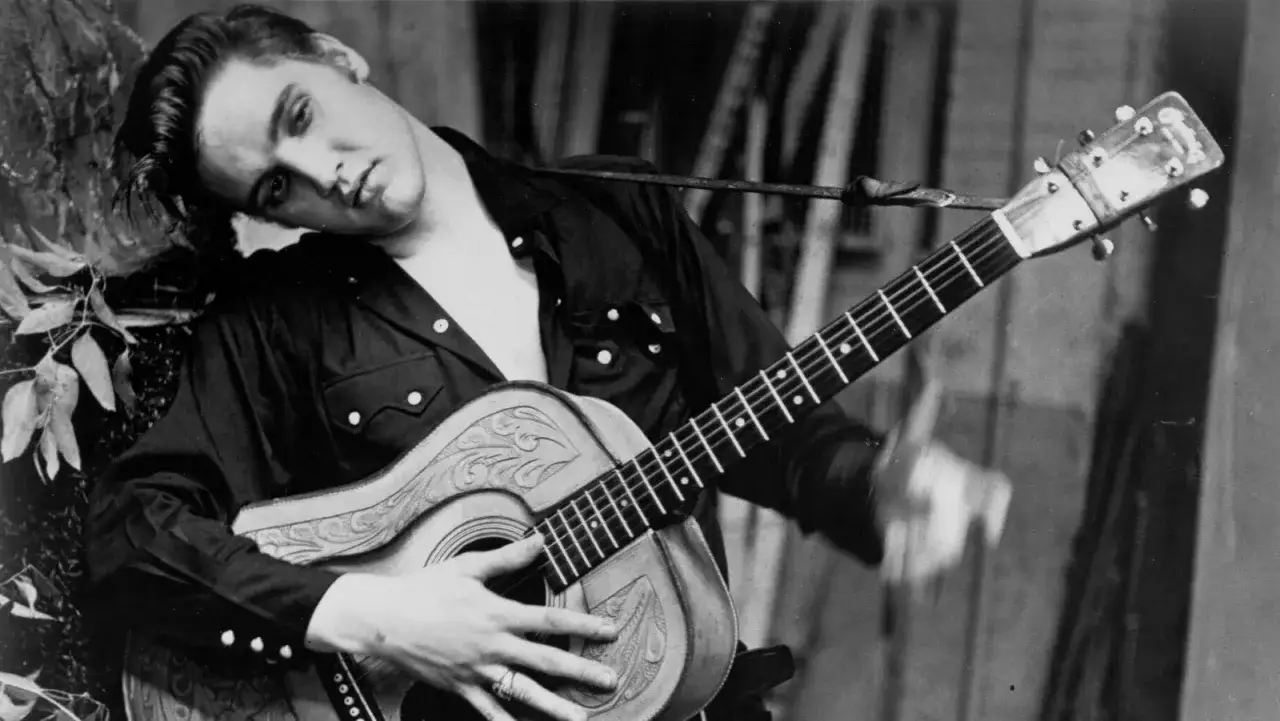 Junger Elvis Presley mit Gitarre, sein Kopf ruht auf seinem Arm, während er sich an einen Baum lehnt.