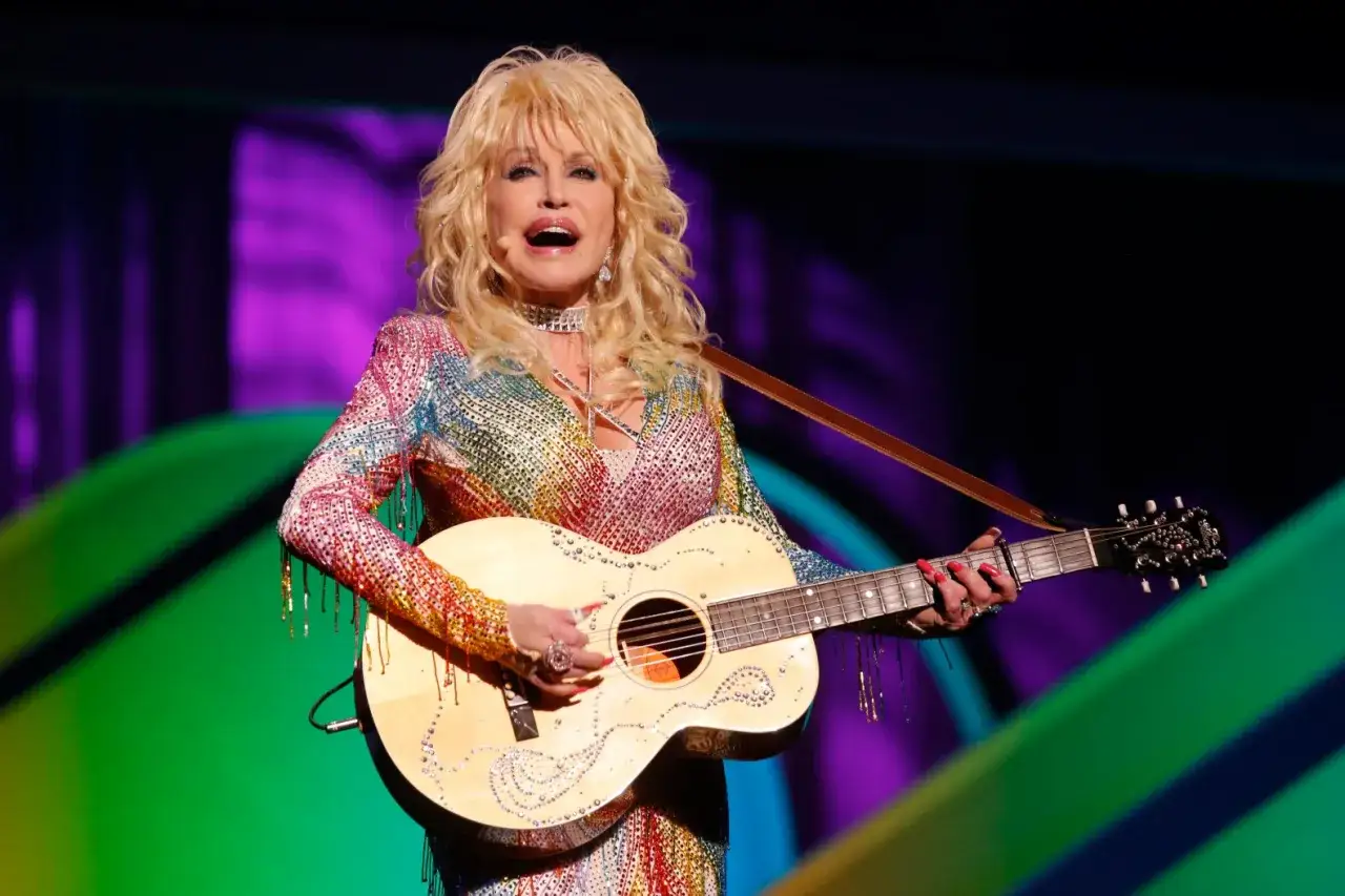 Dolly Parton, die legendäre Country-Sängerin, spielt Gitarre in einem schillernden Regenbogen-Outfit. Sie sieht aus, als wäre sie nie älter geworden, und man fragt sich, wie alt ist Dolly Parton wirklich?