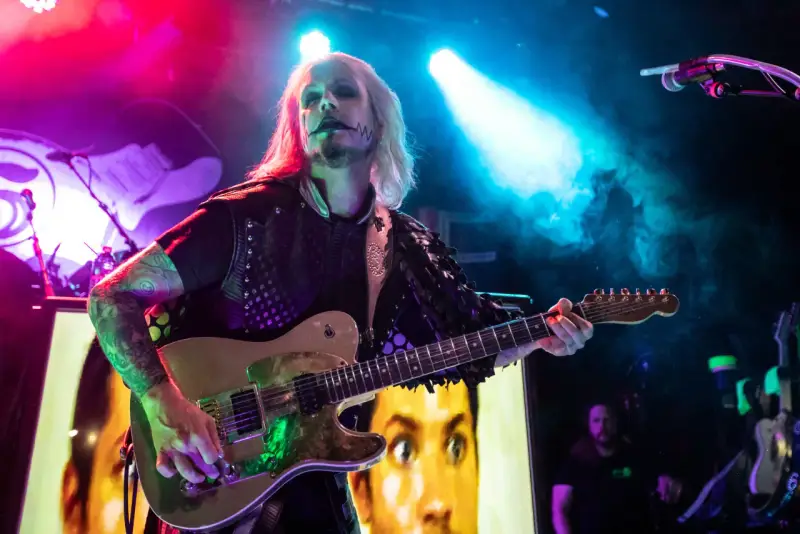 John 5 mit seinem charakteristischen Make-up spielt Gitarre, im Hintergrund ein Bild von Marilyn Manson.